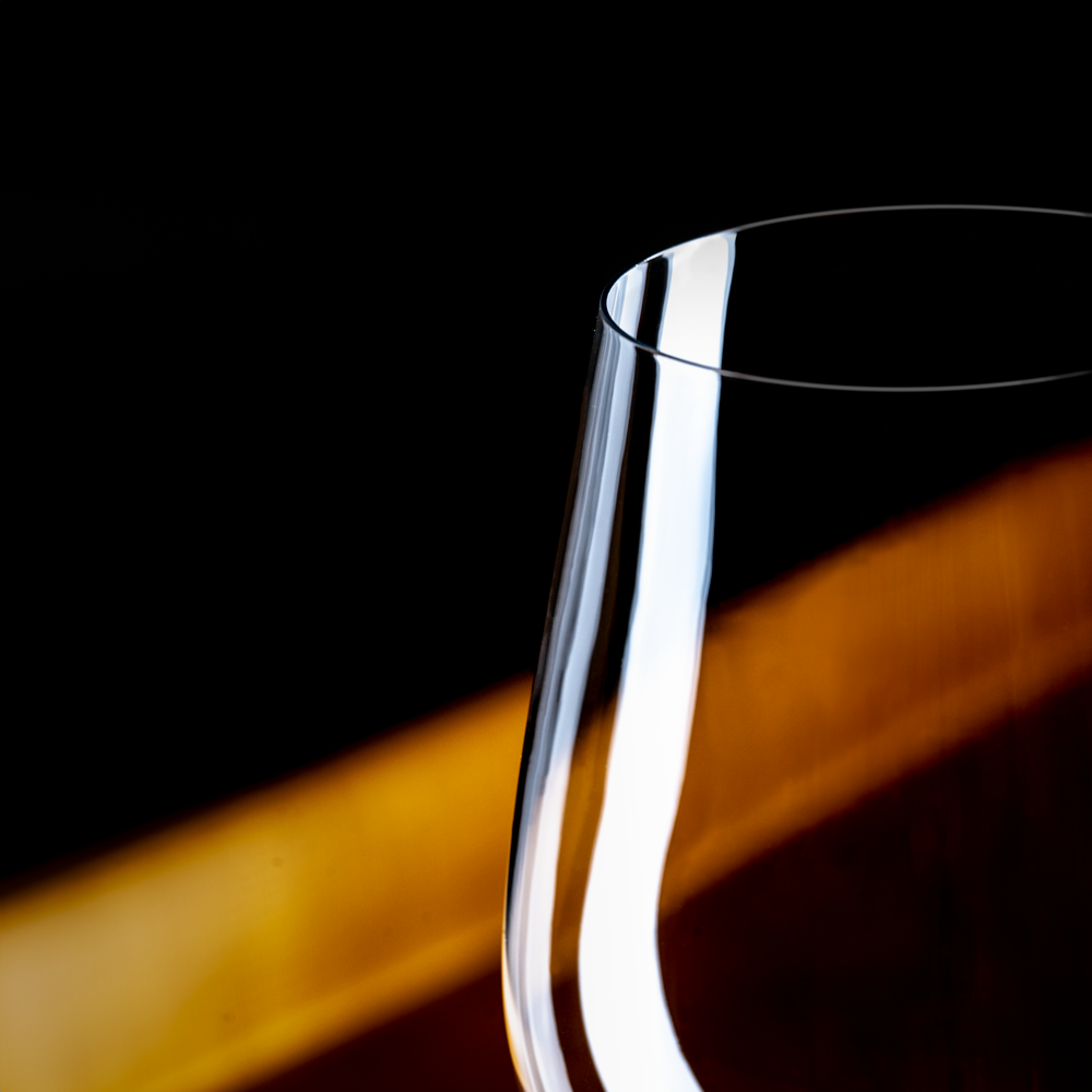 Close-up van de dunne rand van een leeg Lehmann Hommage-glas van 60 cl, tegen een donkere achtergrond met een strook warm licht
