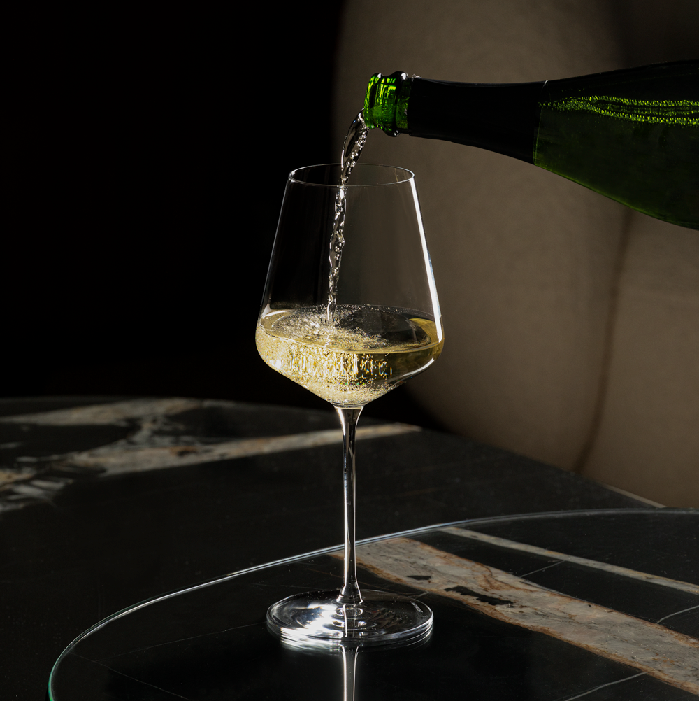 Lehmann Bacchus 55 waarin mousserende wijn uit een groene fles wordt geschonken, op een tafeltje van donker marmer voor een beige bank