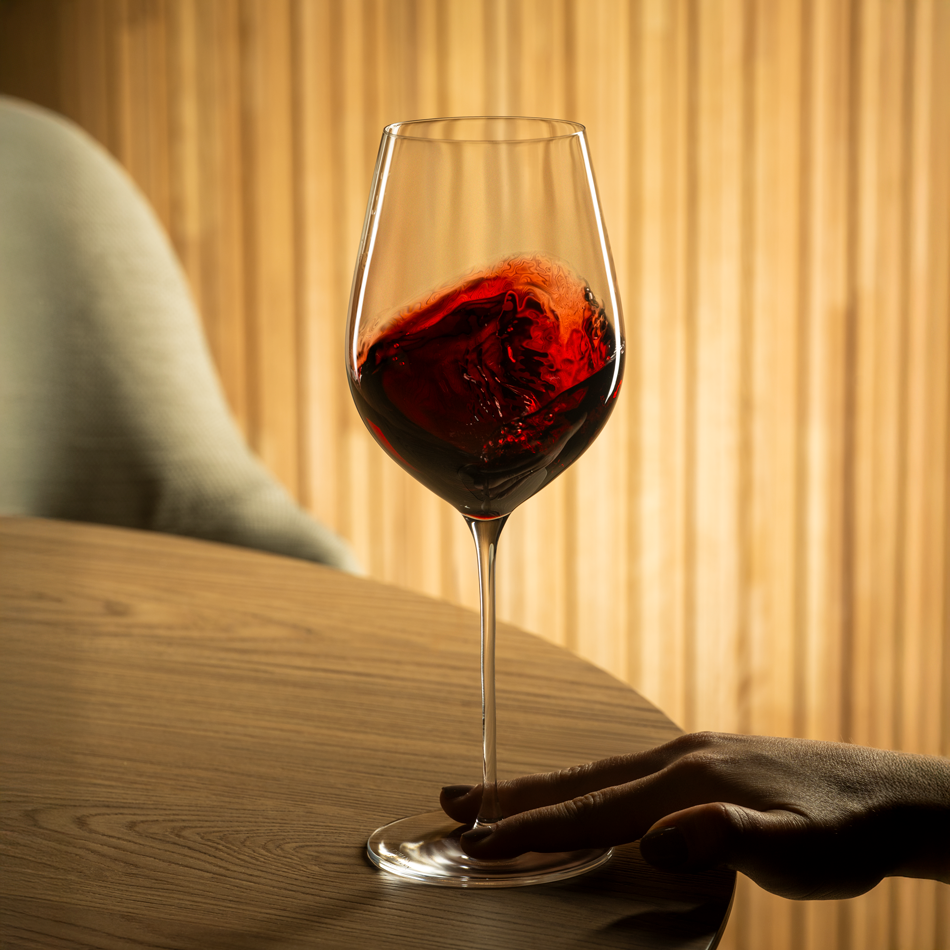Een hand houdt een Lehmann Hommage-glas van 60 cl aan de voet vast op een houten tafel, terwijl de rode wijn erin ronddraait; op de achtergrond een wand van latten