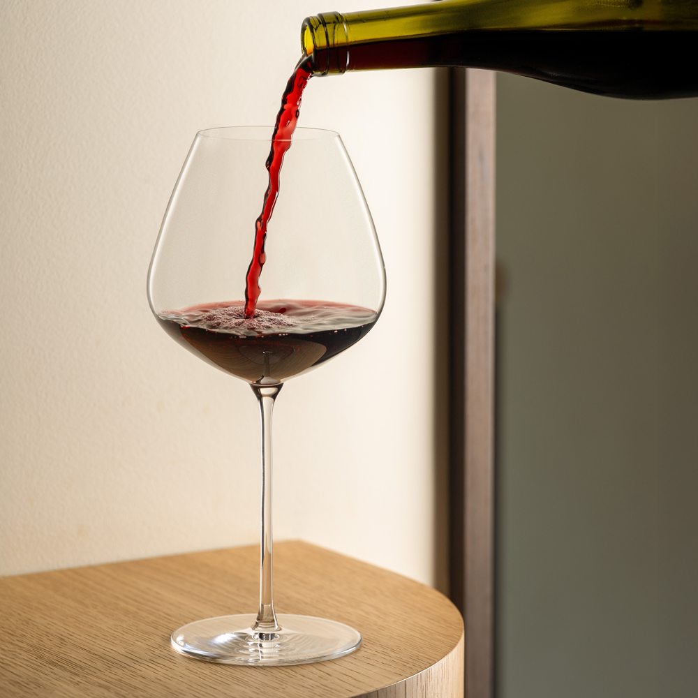 Rode wijn wordt uit een fles in een Lehmann Hommage-glas van 72 cl geschonken, op een houten bijzettafel voor een lichte muur