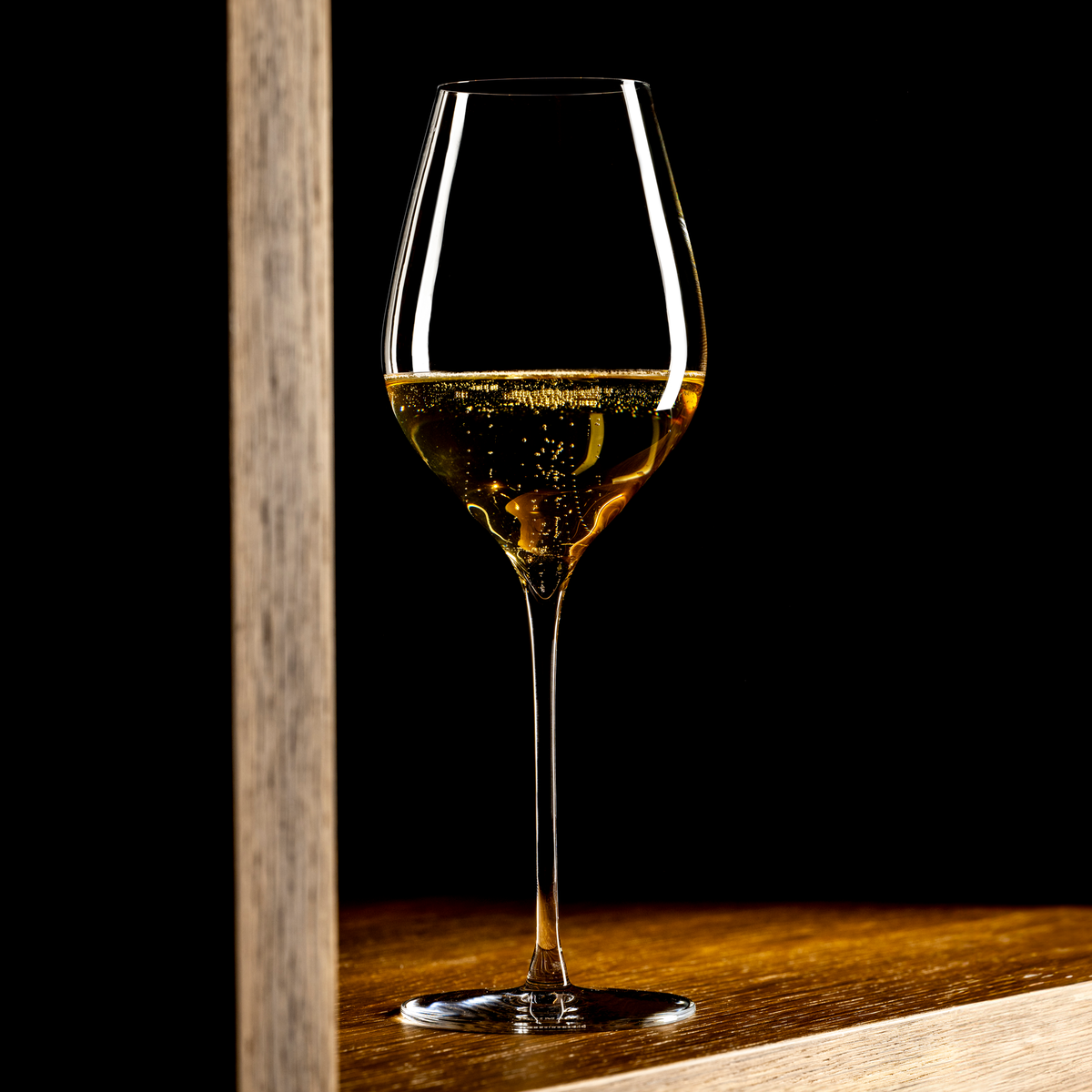 Lehmann N°4 40 Ultralight, half gevuld met goudgele champagne, op een houten blad naast een lichte houten staander, tegen een zwarte achtergrond