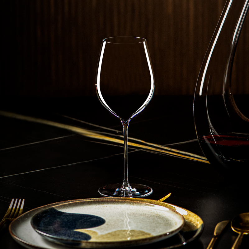 Leeg Lehmann Hommage-glas van 60 cl op een gedekte tafel met een keramisch bord en goudkleurig bestek, rechts een karaf met rode wijn
