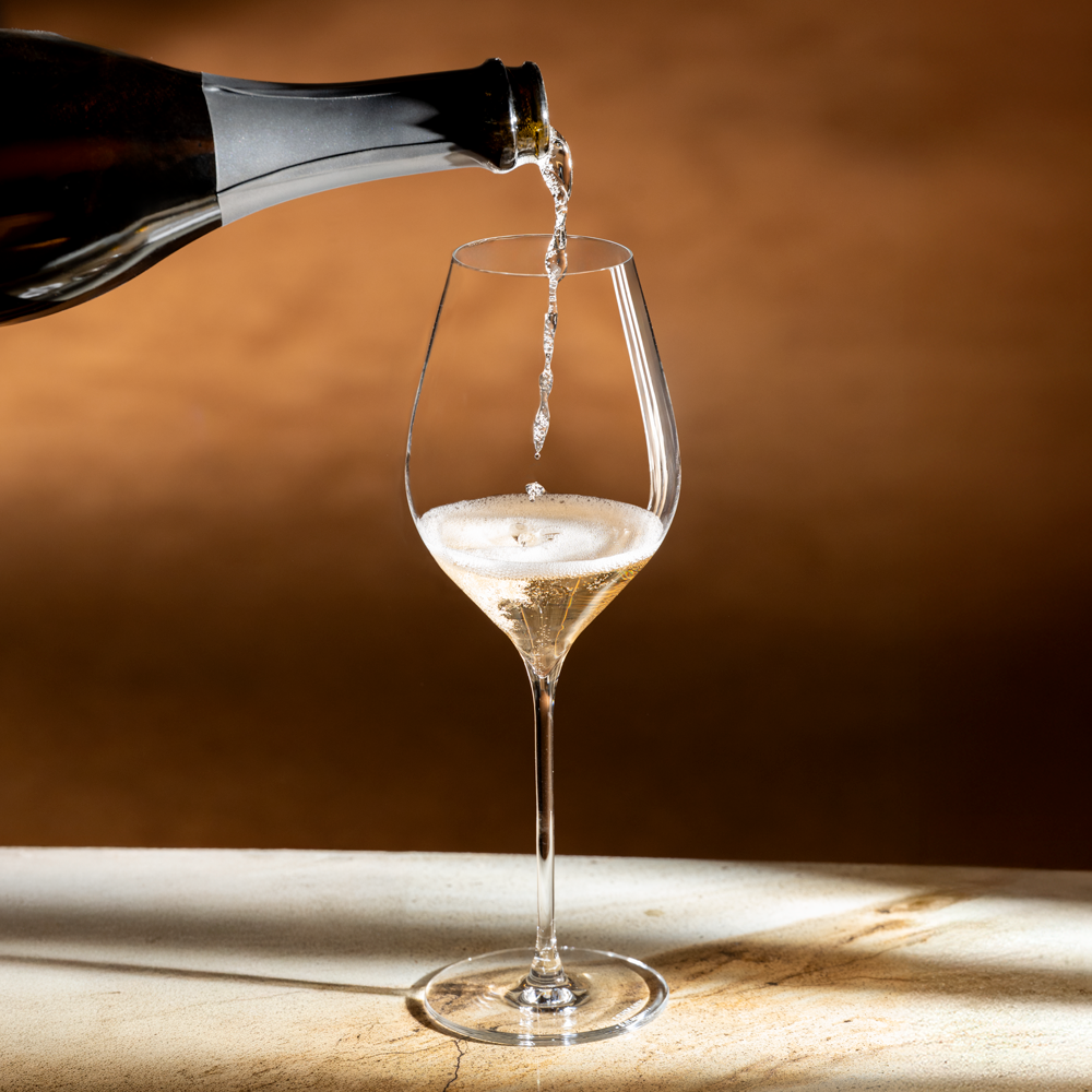 Champagne wordt uit een fles in een Lehmann N°4 40 Ultralight geschonken, op een licht stenen blad voor een warmbruine muur in strijklicht